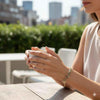Woman wearing a delicate gemstone bracelet holding a cup outdoors, modern city background